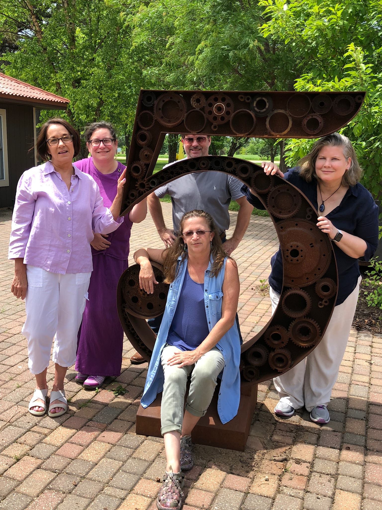 Group photo with the metal “5” at the north studio patio (five studios in this area). [Image description: five Deaf artists are posing around the metal “5” sculpture. Brenda is on the left in a lavender shirt and white pants with lavender fingernails. To the right of Brenda is Lilah, dressed in purple cotton. Behind the 5 is Tony, arms akimbo. Sitting below Tony on the 5 is Cynthia in denim sleeveless long shirt with tan capri pants and blue tank top. To the far right is Katherine, in a black V neck shirt with a pretty necklace and tan pants. All are smiling].