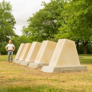 Five large, limestone trapezoids increasing in size in a grassy sculpture garden with trees in background. Sculptor Pete Driessen stands behind his work.