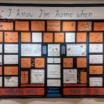A quilt in orange, black, and white with blue accents is made up of rectangular blocks with embroidered text. It reads: I know I'm Home When...