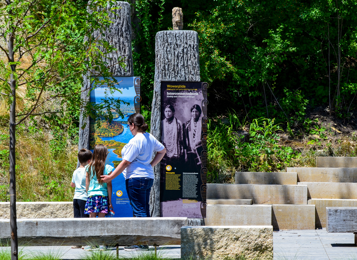 An adult with long hair and two children read interpretive text on large, figure-like, full-color panels. Visible text reads "Wowanzida , Interconnectedness."
