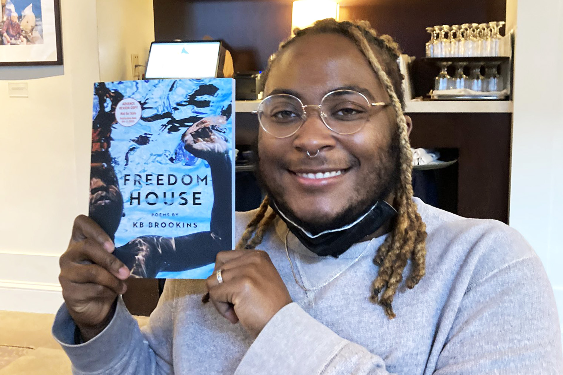 KB — a Black person with brown skin and blond braided locs -- proudly holds up their new book "Freedom House." KB is wearing a gray sweatshirt and round frame glasses. Location is in a residential setting. They are looking directly at the camera.
