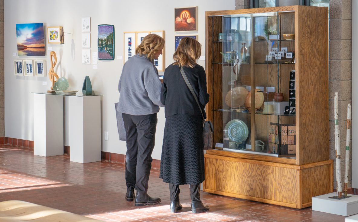 Two women view paintings and ceramic artwork in a gallery.