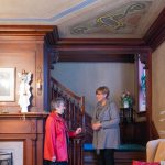 Two women talk within a historic home that has gorgeous woodwork and original decorative painting on the walls