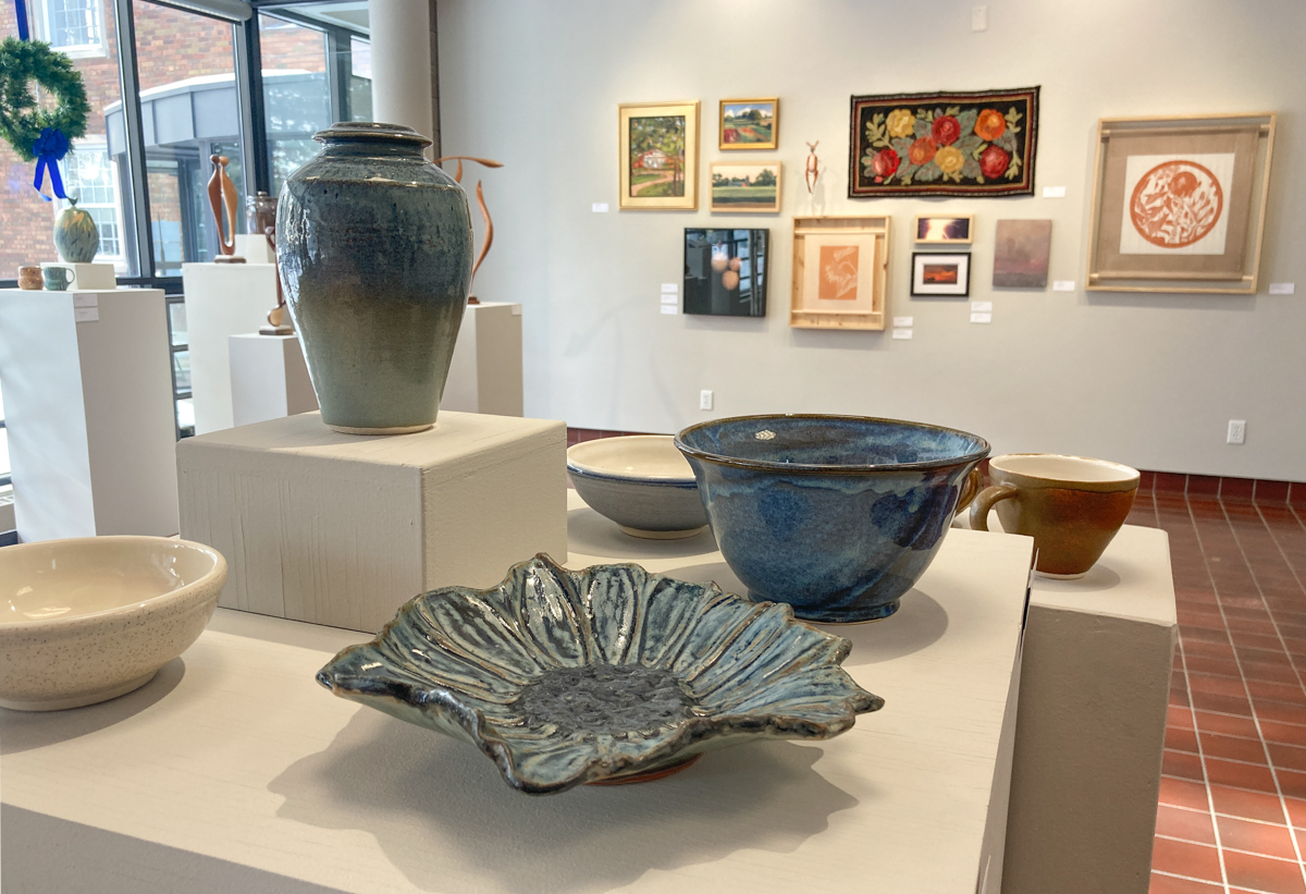 ceramic bowls and vases sit on pedestals in the foreground with a variety of 2-dimensional art on the wall in the background.