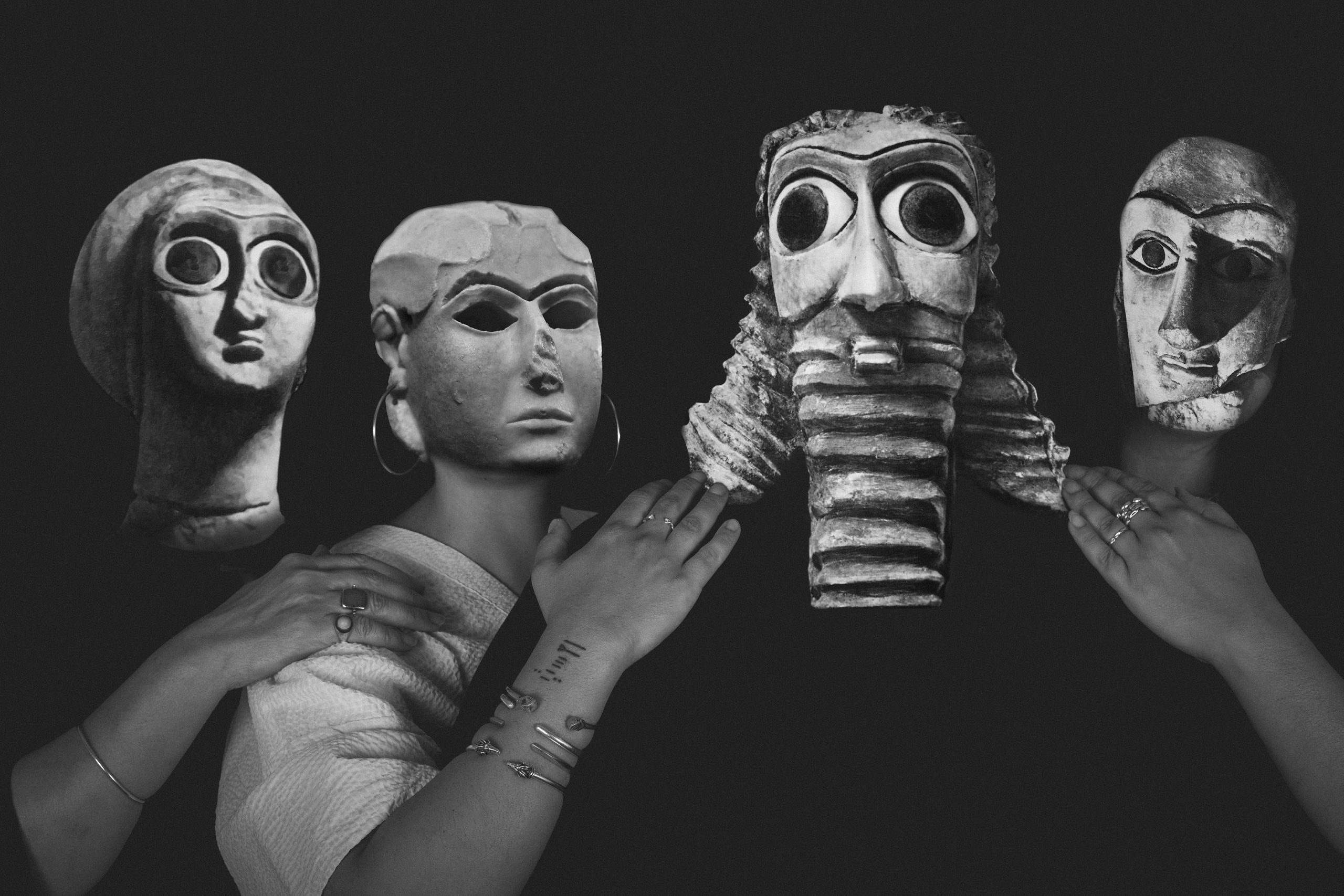 A black-and-white photograph shows four figures from the bust up. They are wearing masks that reference ancient Sumerian artworks and have their hands on each others shoulders.