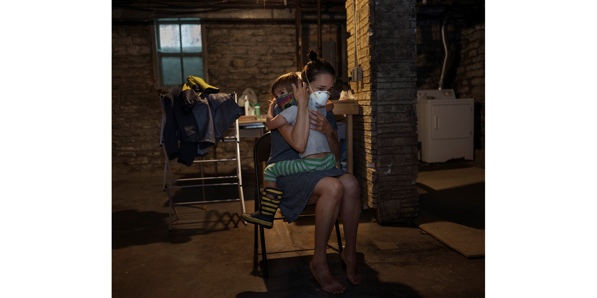 A woman with dark hair holds a young boy with light hair. They are in a basement space and both wear face masks.