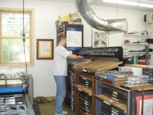 A man in a white t-shirt and jeans sets type in a studio surrounded by type drawers