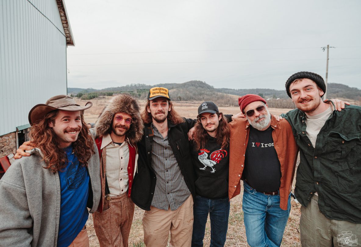 Six members of Saltydog pose outside against a steel building and a hilly setting