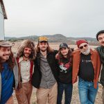 Six members of Saltydog pose outside against a steel building and a hilly setting