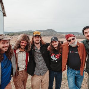 Six members of Saltydog pose outside against a steel building and a hilly setting