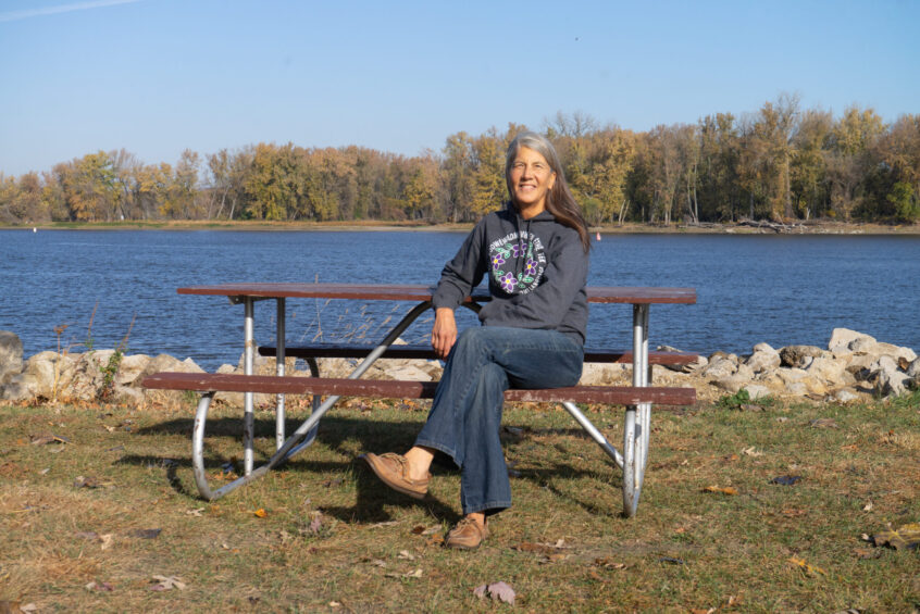 Margaret Pearce sits at a picnic table on the shoreline of the Mississippi River.